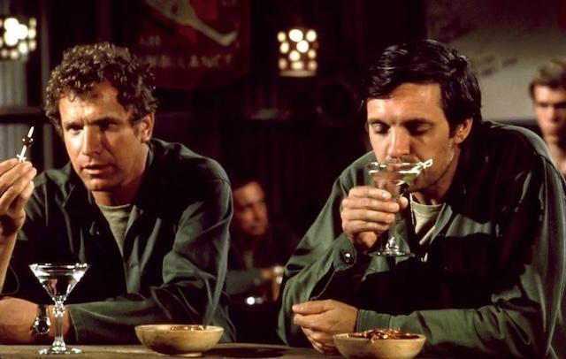 Wayne Rogers as Trapper John and Alan Alda as Hawkeye Pierce sharing a serious conversation over martinis in the iconic Swamp tent from M*A*S*H.